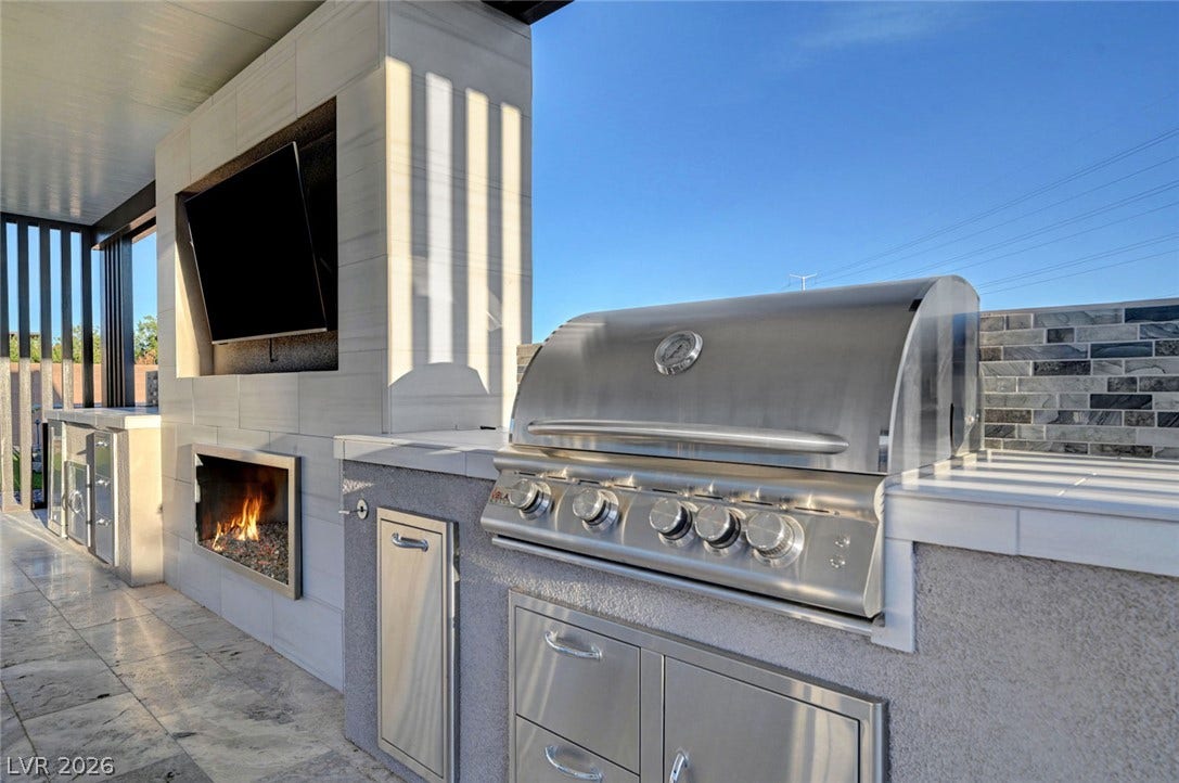 An outdoor kitchen features a built-in stainless steel grill and a modern fireplace.