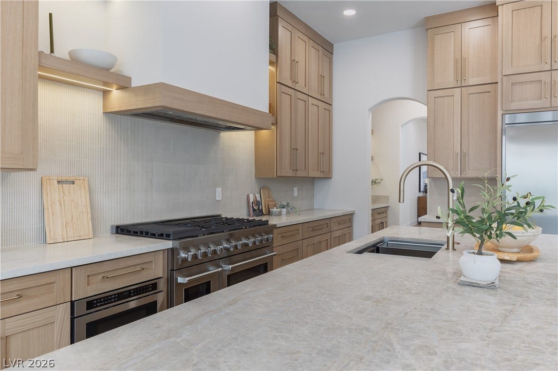 Kitchen featuring high quality appliances, light wood finish cabinetry, light stone countertops, decorative backsplash, and arched walkways