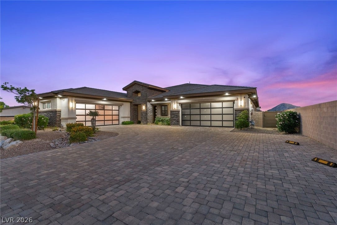 9841 W El Campo Grande Avenue, Las Vegas, NV 89149 A grand paver driveway leads to a modern facade with glass garage doors, illuminated under a twilight sky.