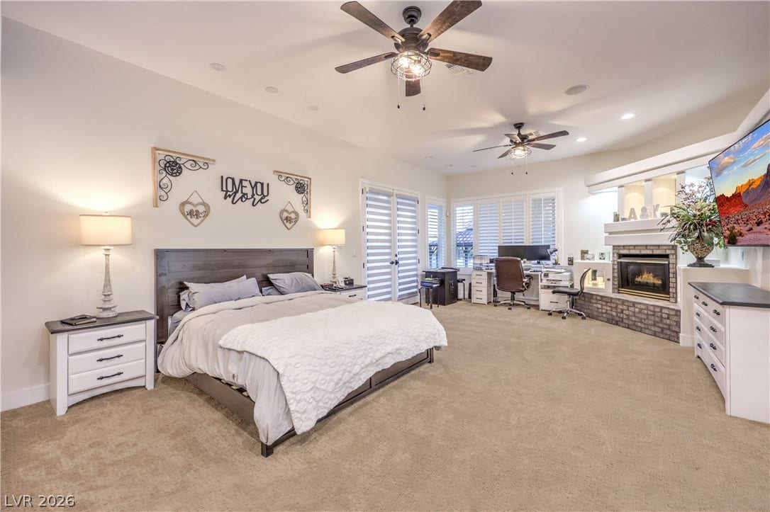 A spacious bedroom suite showcases neutral carpeting, recessed lighting, and a brick-accented fireplace.