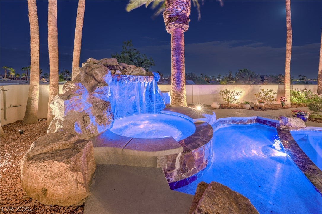 A stone waterfall cascades into the built-in spa beside the swimming pool under palm trees.