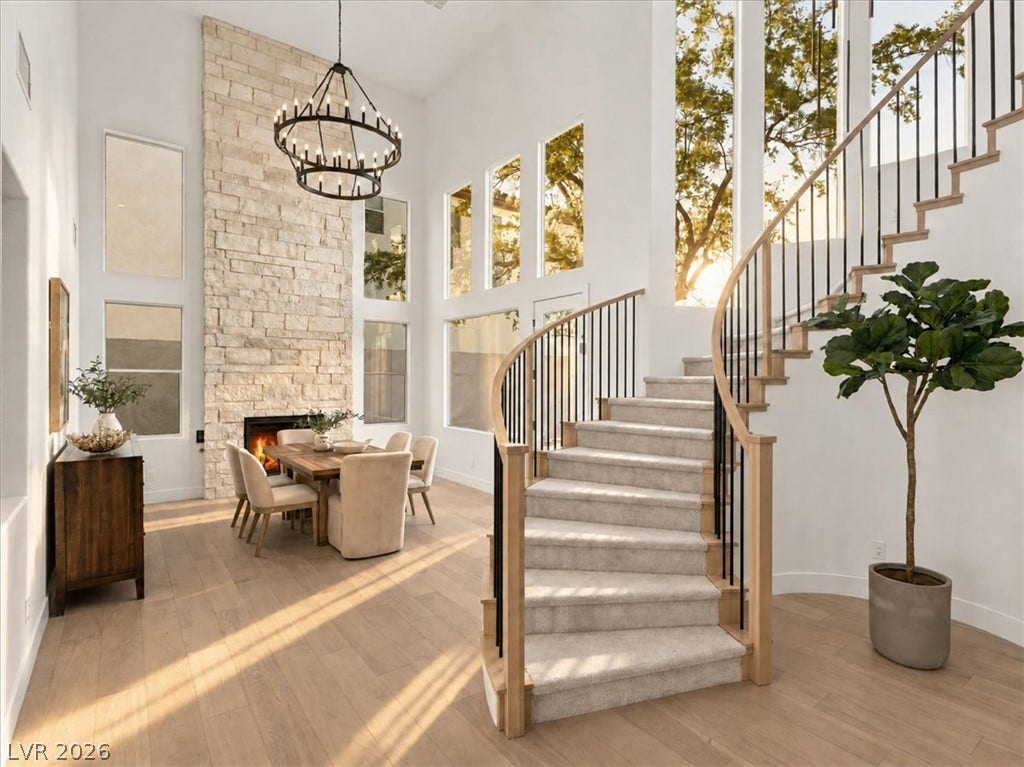 A striking curved staircase ascends alongside a soaring stone-clad fireplace and abundant windows.