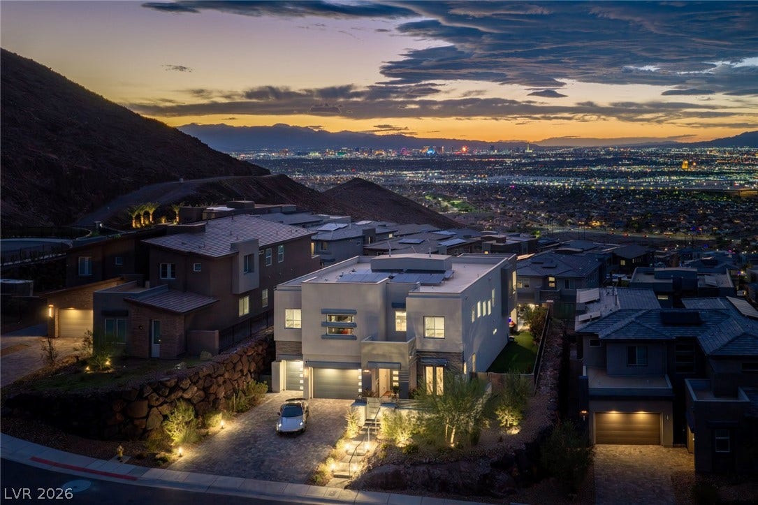 A modern hillside residence overlooks a glittering valley with mountain ridges under a sunset sky.