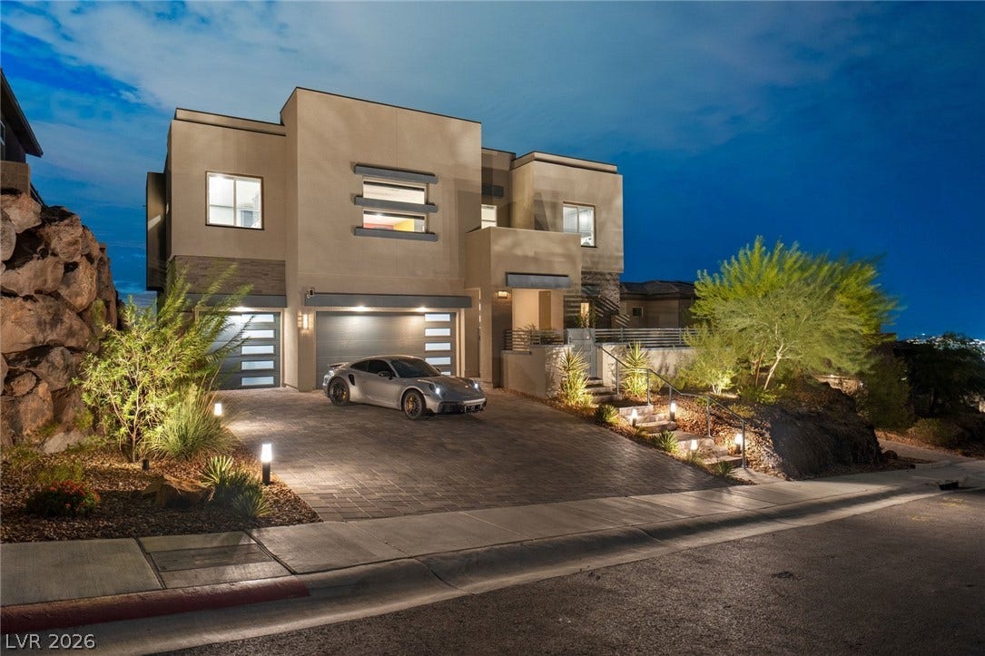 Striking contemporary architecture rises beside a paved driveway highlighted by integrated landscape illumination.