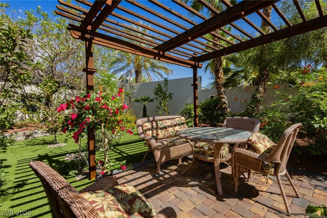 A quiet garden patio sits beneath a classic wooden pergola surrounded by lush greenery.