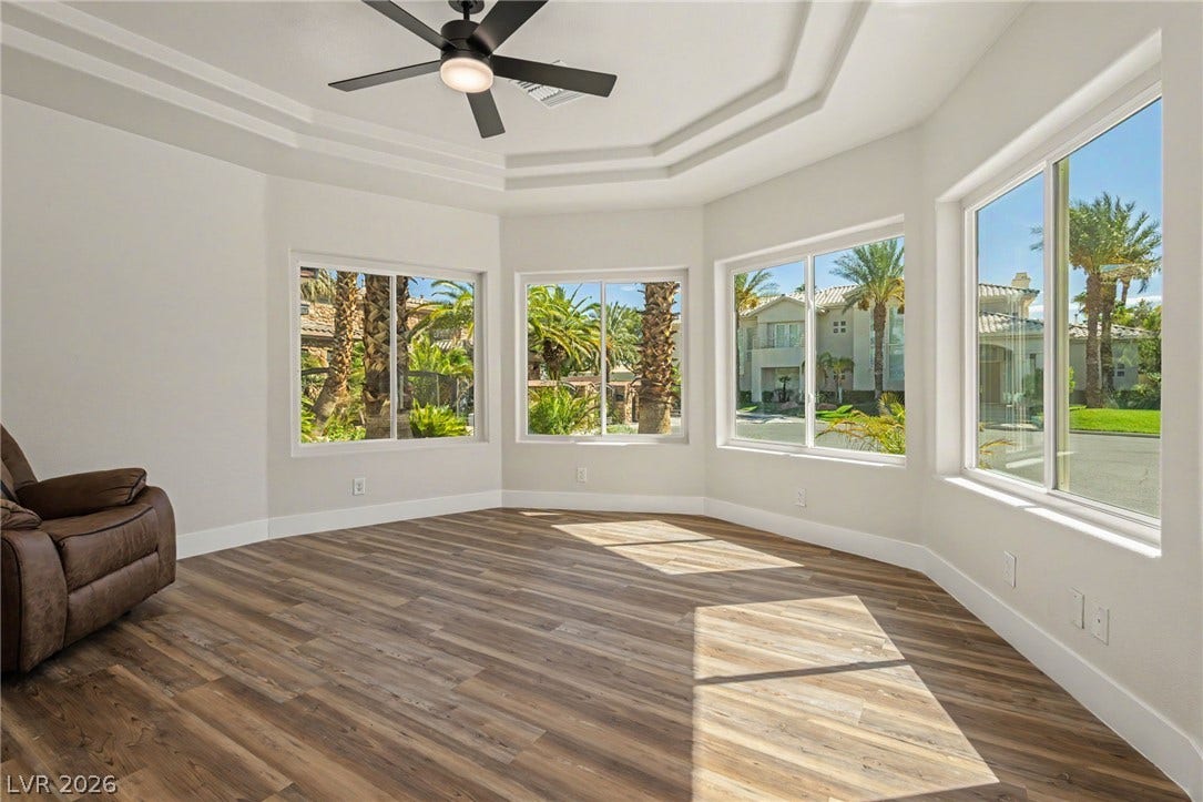 Sunlight streams across wood flooring through a bay of expansive windows under a tiered tray ceiling.