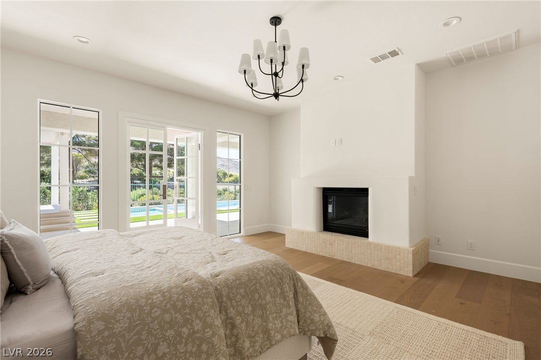 12007 Oakland Hills Drive, Las Vegas, NV 89141 A modern fireplace and classic French doors elevate this light-filled primary suite with hardwood flooring.