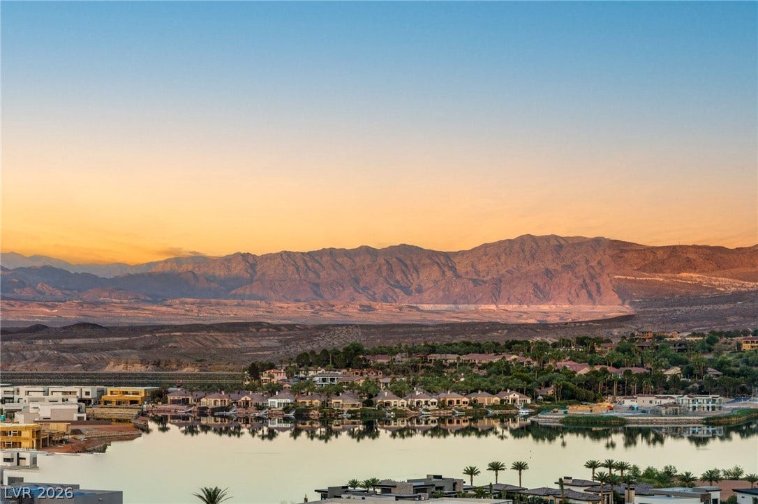 A vibrant sunset over tranquil waters, nestled below majestic mountain ranges and a community of homes.