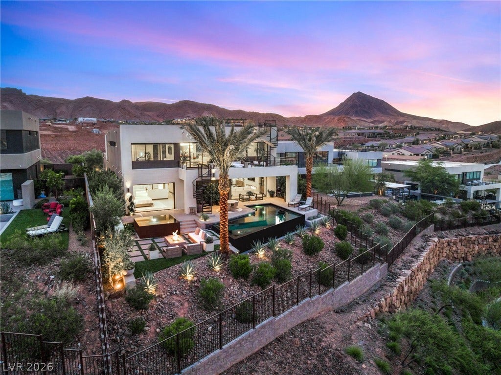 An aerial perspective reveals a luxurious hillside residence with a private pool and illuminated patio areas against a vibrant sunset.