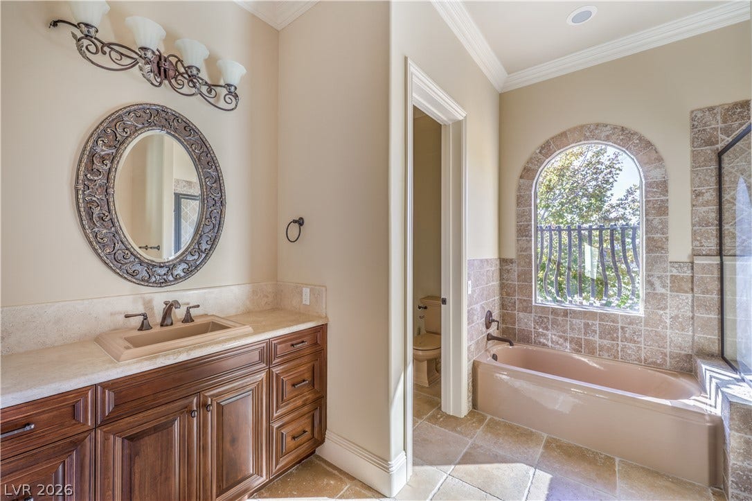 16 Anthem Pointe Court, Henderson, NV 89052 An elegant stone-tiled bathtub rests beneath an arched window framing serene outdoor views.