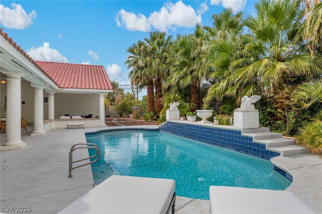 Lush palms surround a serene swimming pool bordered by a tiled feature wall and concrete patio.