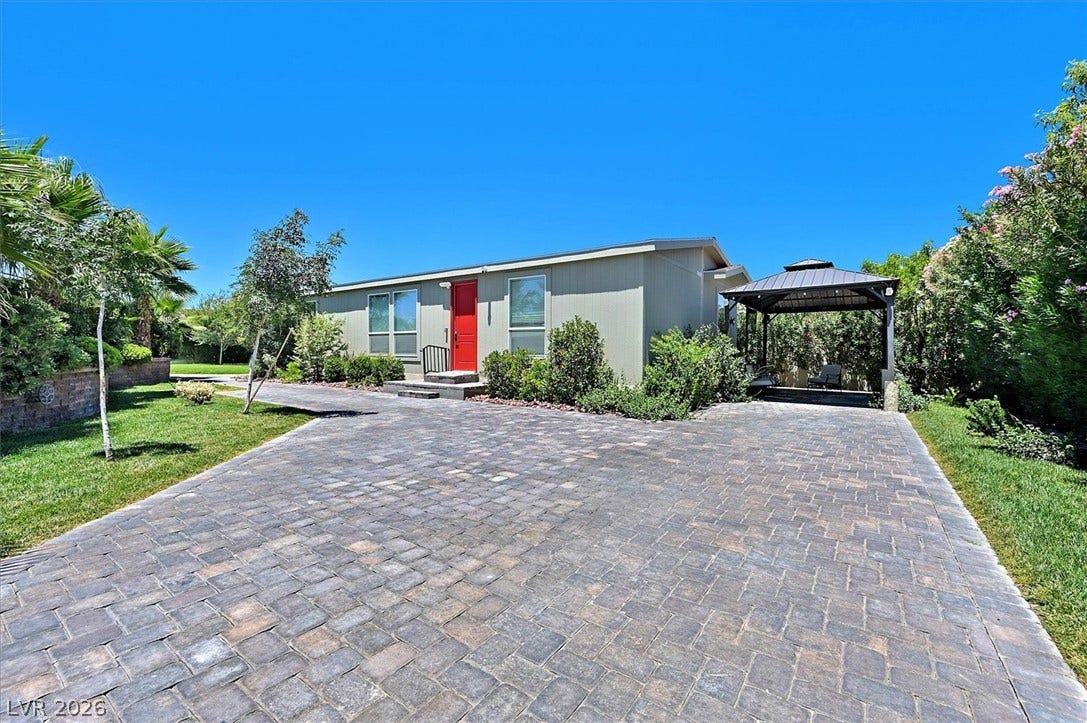 A long paver driveway leads to a modern home with a vibrant red door and a shaded outdoor seating area.