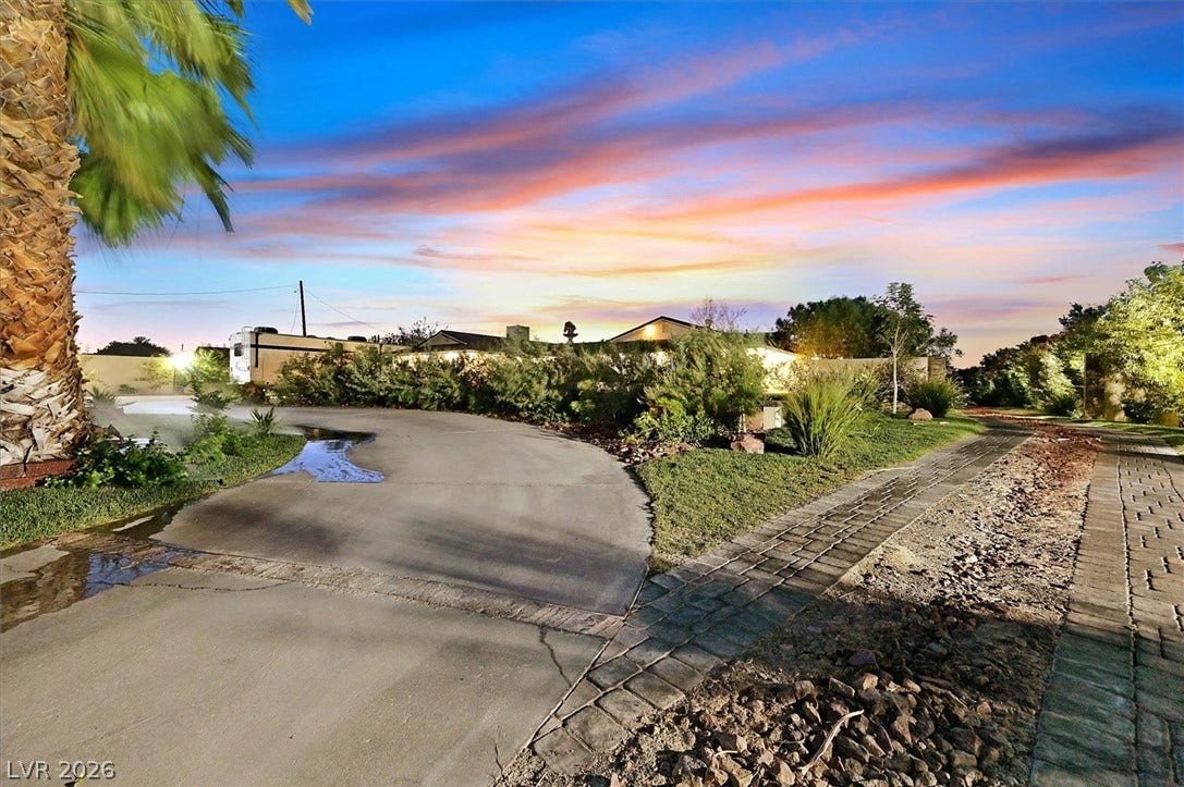Dramatic sunset hues illuminate the spacious paved driveway and mature landscaping, creating an inviting approach.