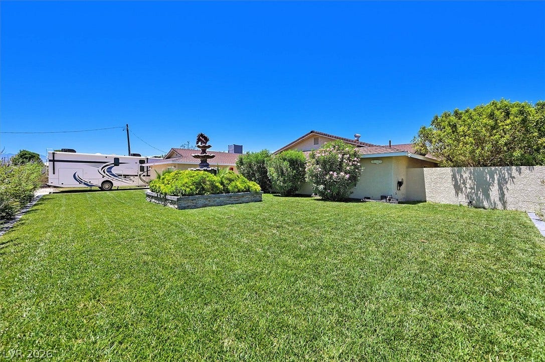 A broad, well-tended green lawn extends toward a stone fountain feature and a distant RV.