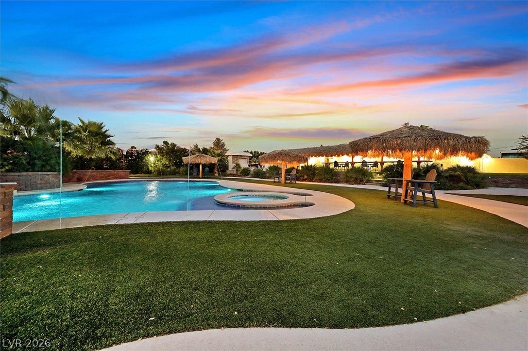 An inviting backyard oasis offers a sparkling pool with an integrated spa, set amidst lush artificial turf and tropical tiki huts.