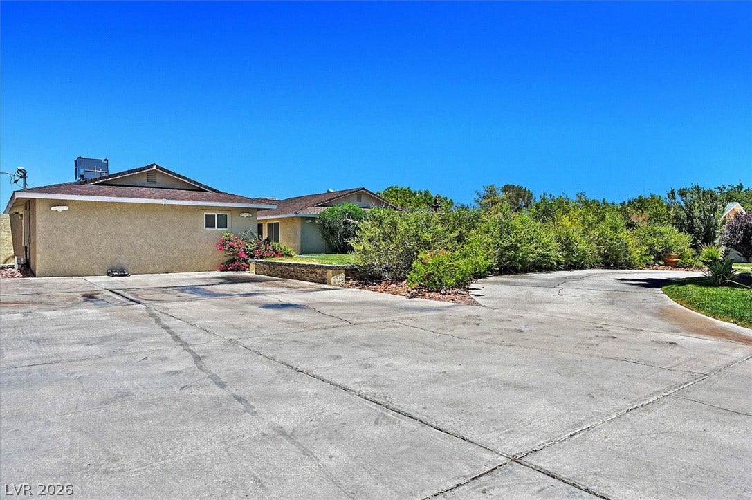 Concrete driveway leads to a single-story residence with a stucco exterior and surrounding greenery.