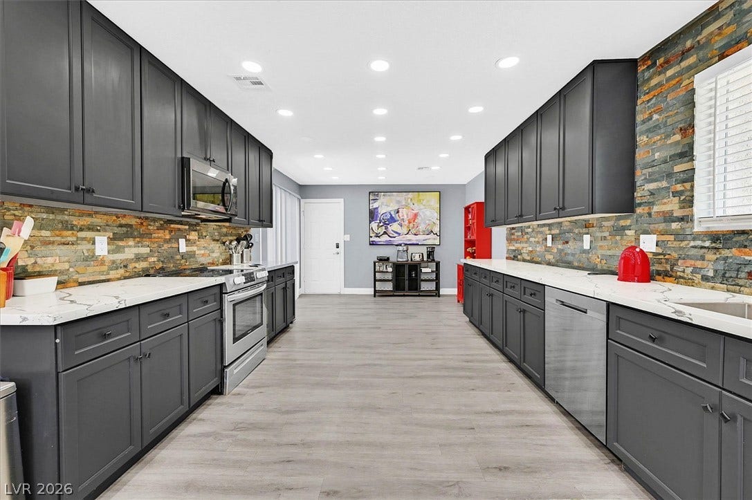 A galley kitchen boasts sleek gray cabinetry and stainless steel appliances against a captivating stacked stone backsplash.