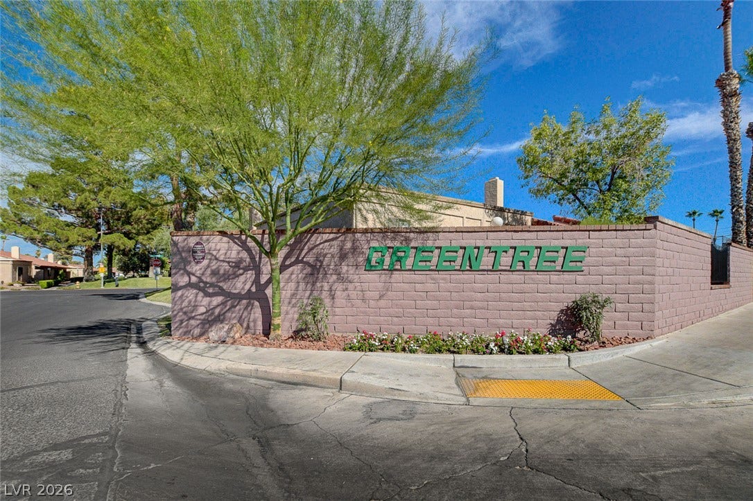 Mature trees shade the brick entry monument bordering wide suburban streets under clear skies.