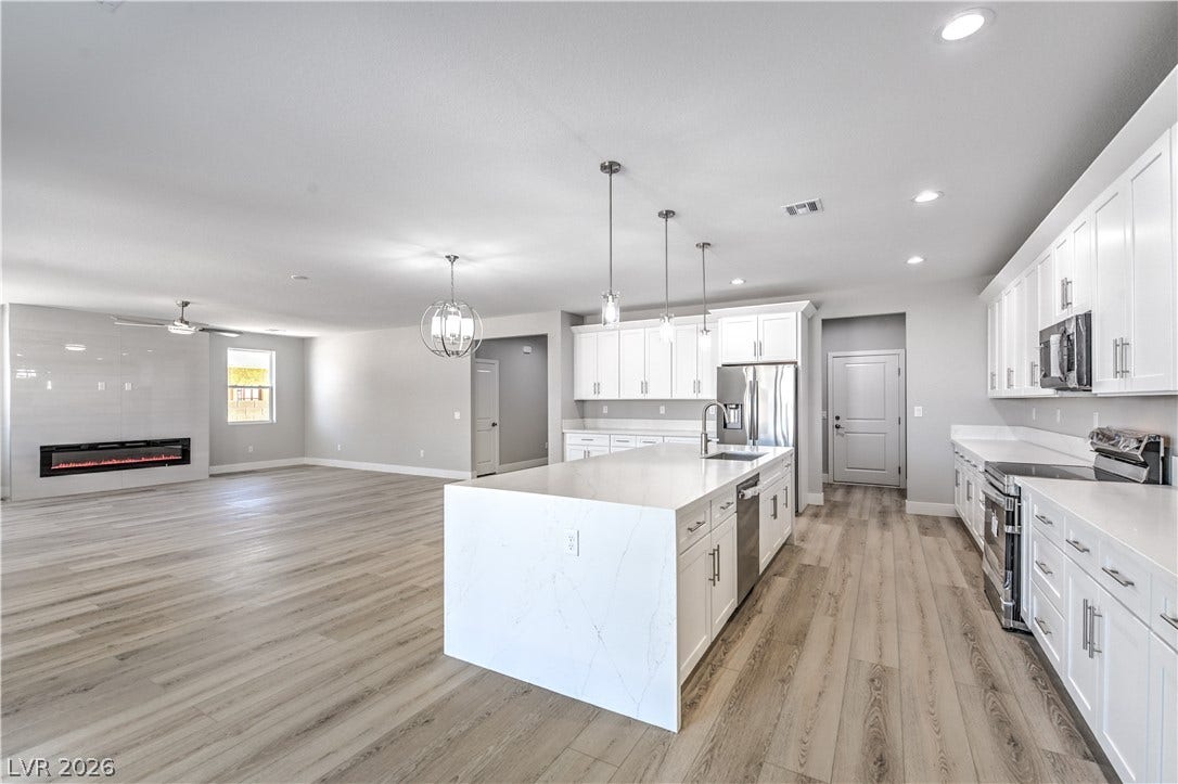Kitchen featuring stainless steel appliances, an island with sink, white cabinetry, light stone countertops, and decorative light fixtures