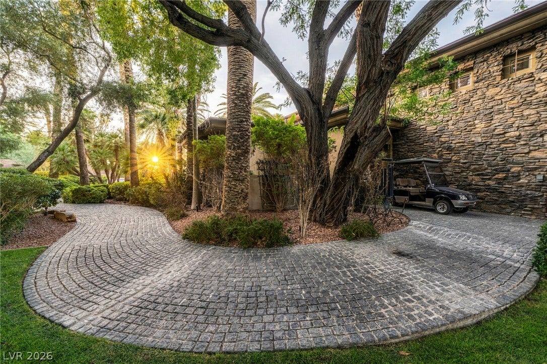 1513 Foothills Village Drive, Henderson, NV 89012 A curved stone paver driveway winds past mature trees toward the rustic stone exterior.