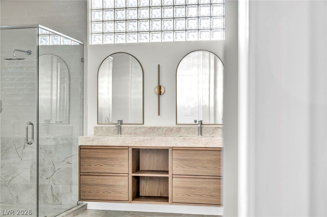 8805 Spanish Mountain Drive, Las Vegas, NV 89148 Arched mirrors and stone countertops elevate this modern double vanity area beside a glass-enclosed shower.