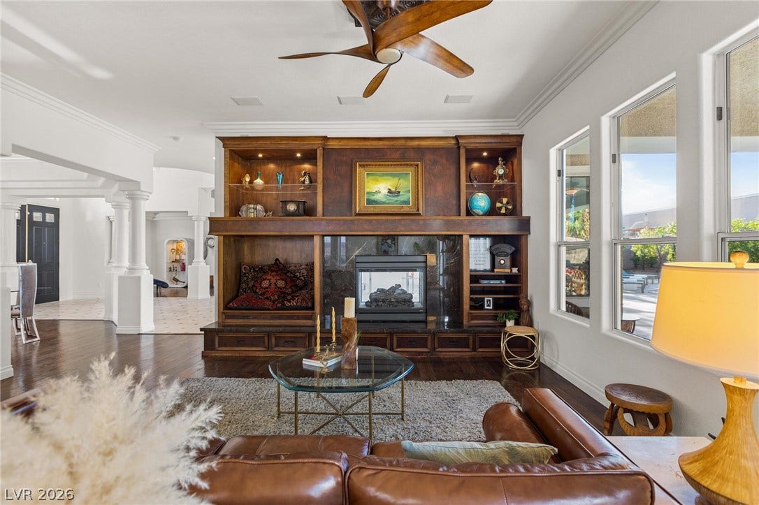 8088 Dark Hollow Place, Las Vegas, NV 89117 A spacious living area boasts a grand fireplace flanked by custom built-in shelving, complemented by a large ceiling fan for ultimate comfort.