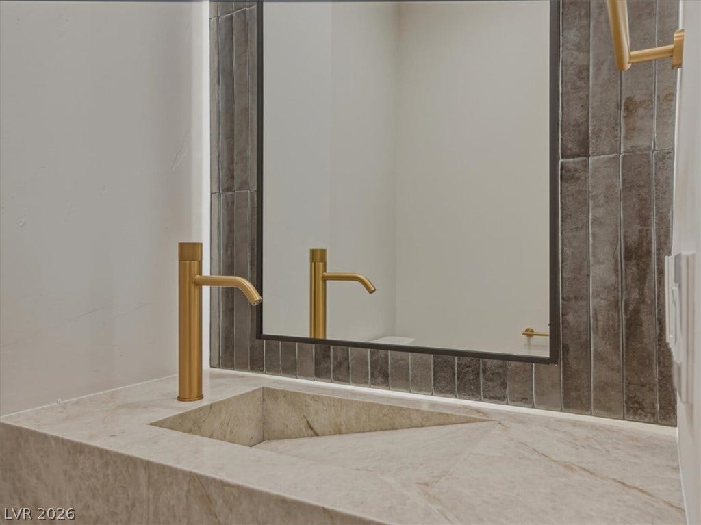 3 Isleworth Drive, Henderson, NV 89052 A modern bathroom vanity showcases a sleek integrated sink and striking gold-toned faucet against a textured tiled mirror frame.