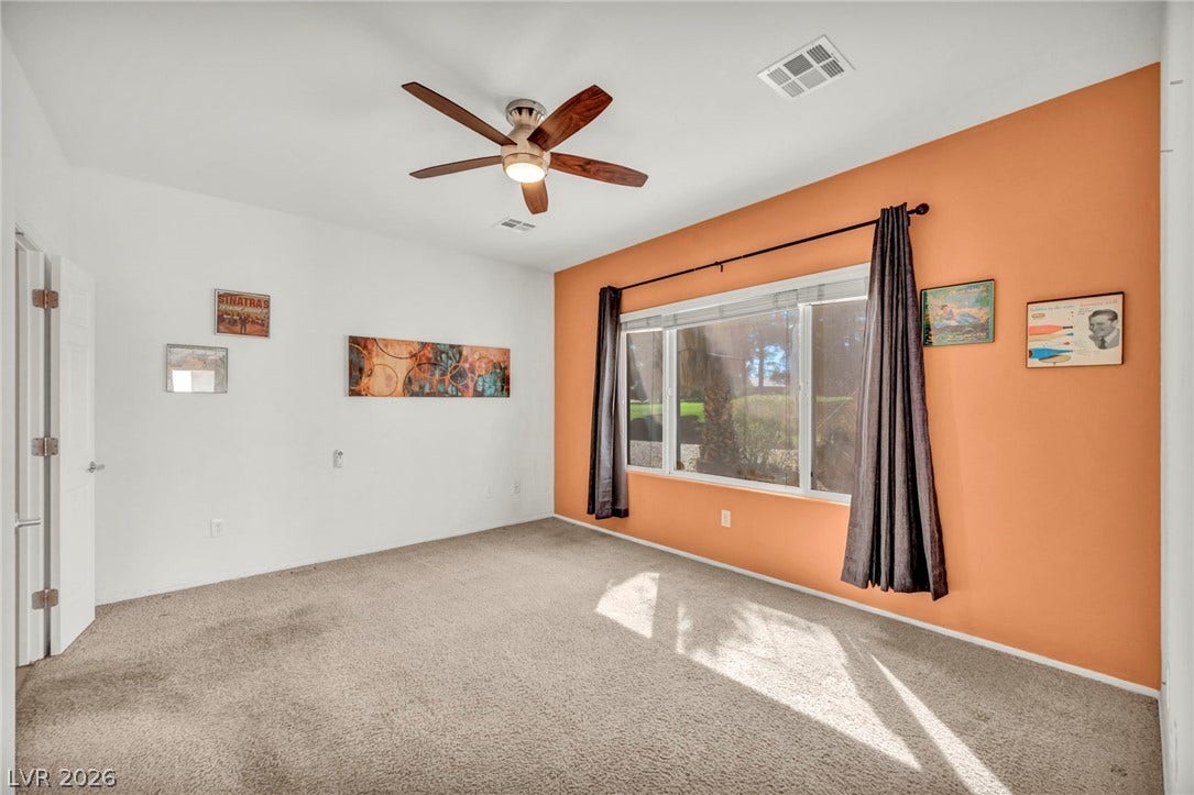 A wide window illuminates the carpeted floor and tall ceiling of this bedroom space.