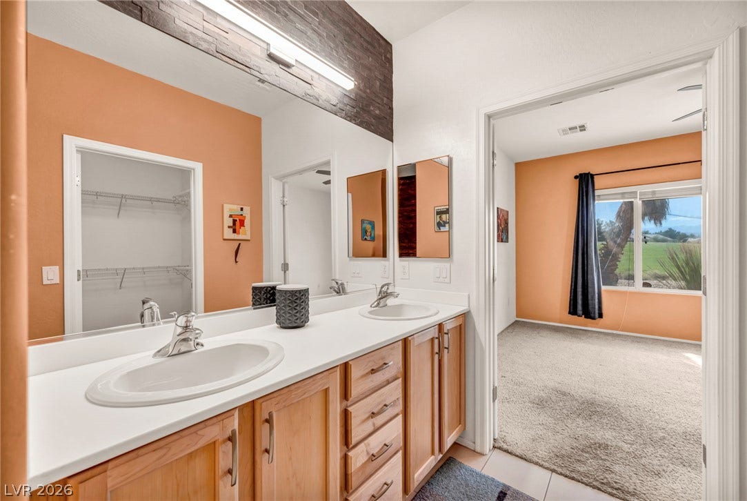 Double vanity sinks and clean cabinetry connect seamlessly to an adjacent carpeted suite.