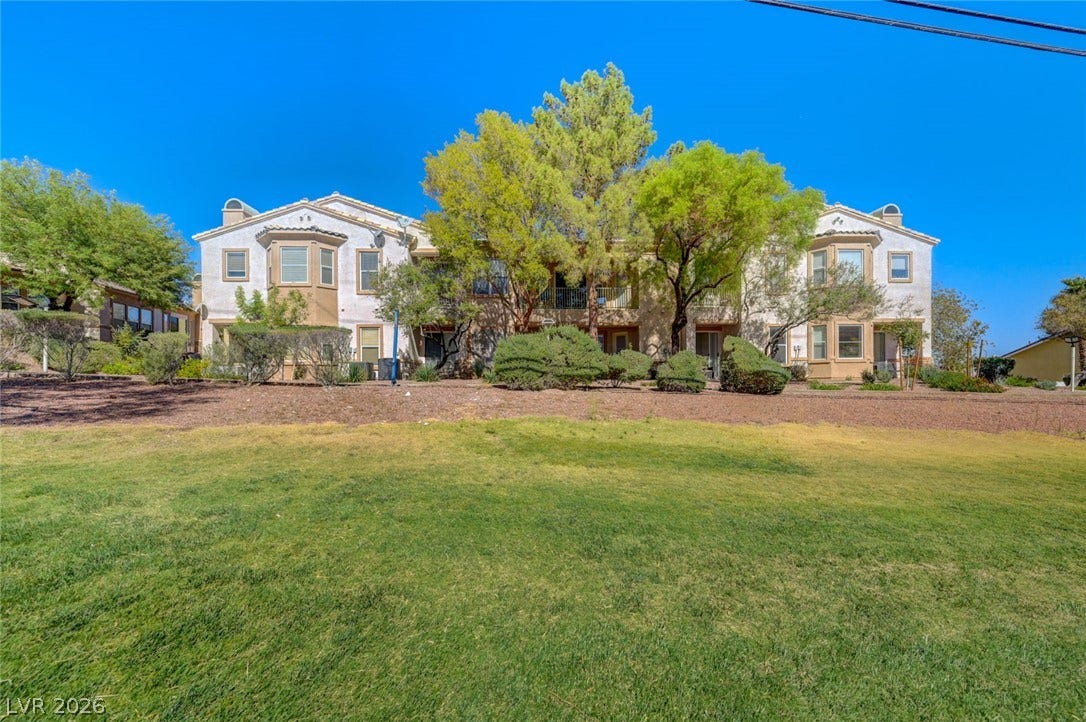 207 Kaelyn Street, #5, Boulder City, NV 89005 Mature trees frame the multi-story stucco facade beyond a wide expanse of green lawn.