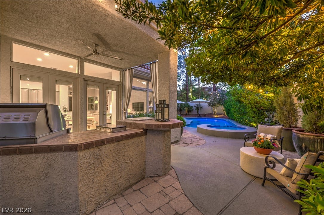Mature trees frame a concrete patio featuring an outdoor kitchen alongside a refreshing backyard pool.