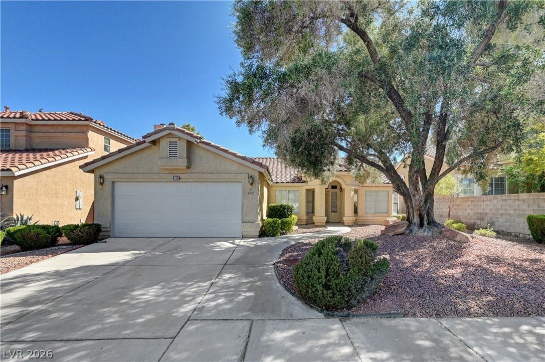 Mature trees and a concrete driveway frame this classic stucco home under clear blue skies.