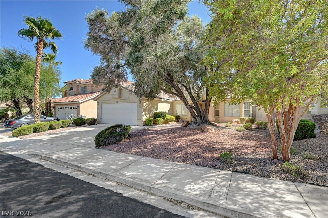 Mature shade trees and desert landscaping frame the concrete driveway leading to a two-car garage.