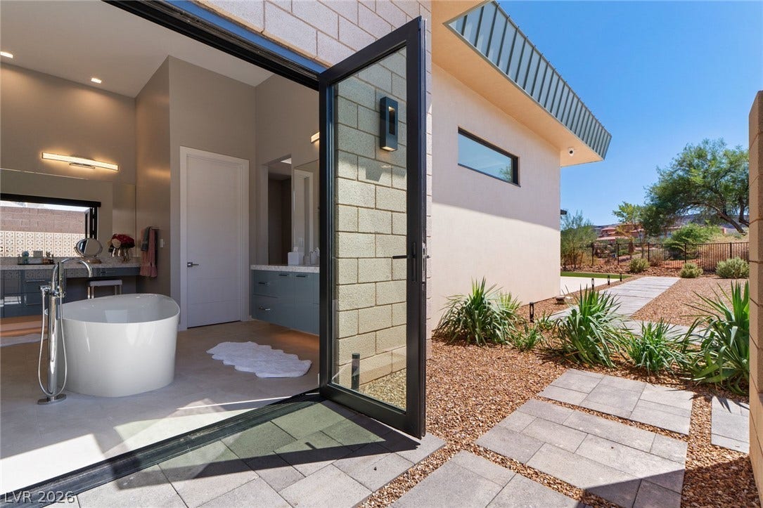 37 Reflection Shores Lane, Henderson, NV 89011 An open glass door connects a modern bathroom featuring a freestanding tub to paved outdoor walkways.