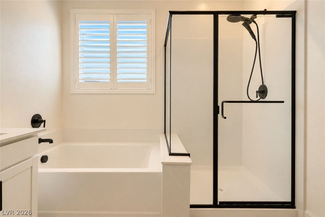 A glass-enclosed walk-in shower stands beside a deep soaking tub below plantation-shuttered windows.
