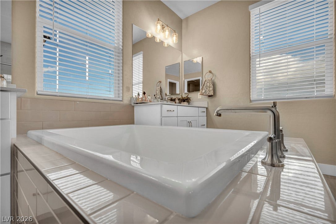 7128 Port Stephens Street, North Las Vegas, NV 89084 A deep soaking tub sits framed by large windows in this modern primary bathroom.