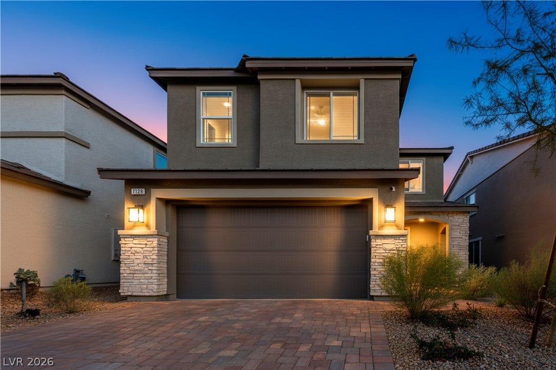 7128 Port Stephens Street, North Las Vegas, NV 89084 Warm exterior lights illuminate the stone accents and paved driveway of this modern two-story home.