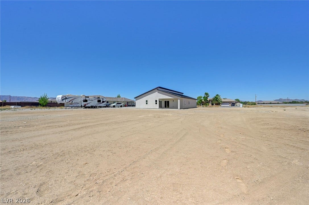 A minimalist stucco exterior stands set back on a vast desert lot under blue skies.