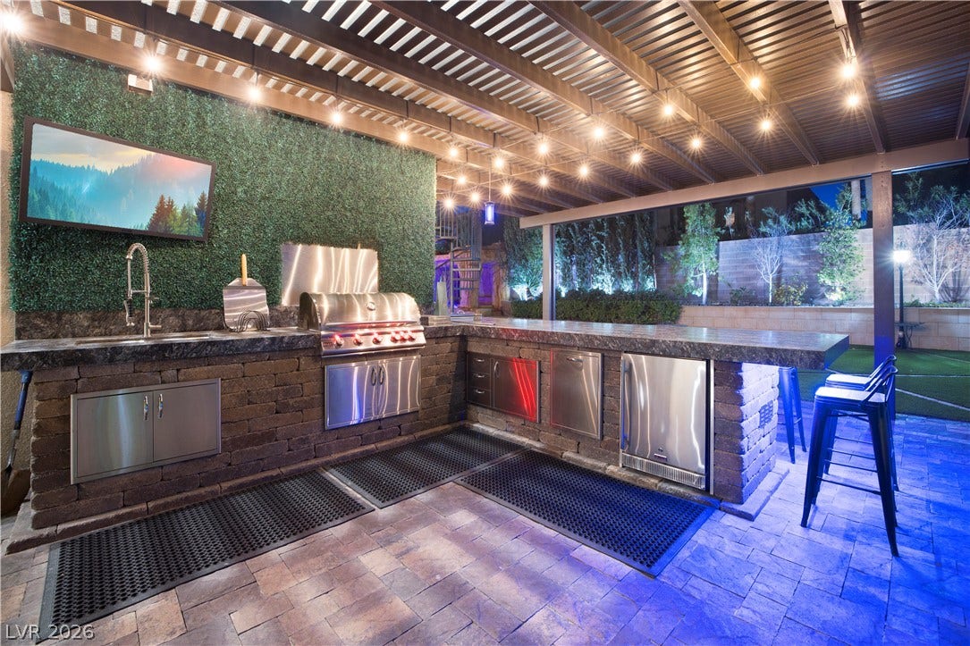 9624 Grand Isle Lane, Las Vegas, NV 89144 An outdoor kitchen sits under a covered pavilion featuring stone pavers and integrated lighting.