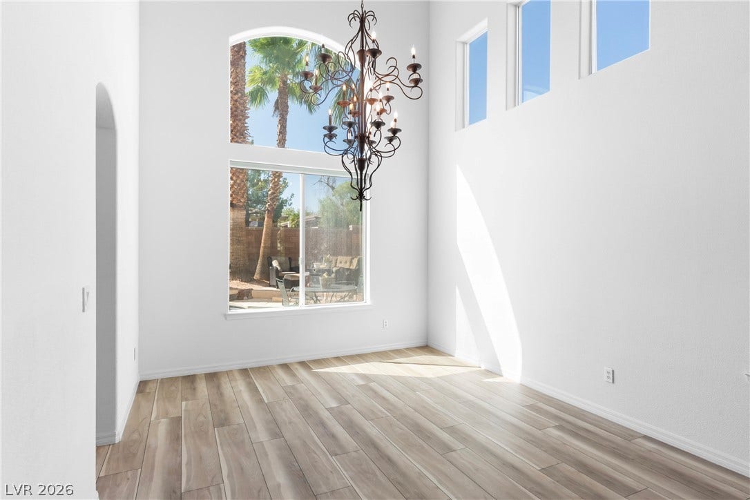 A spacious room features soaring ceilings, a dramatic chandelier, and sun-filled windows overlooking palm trees.