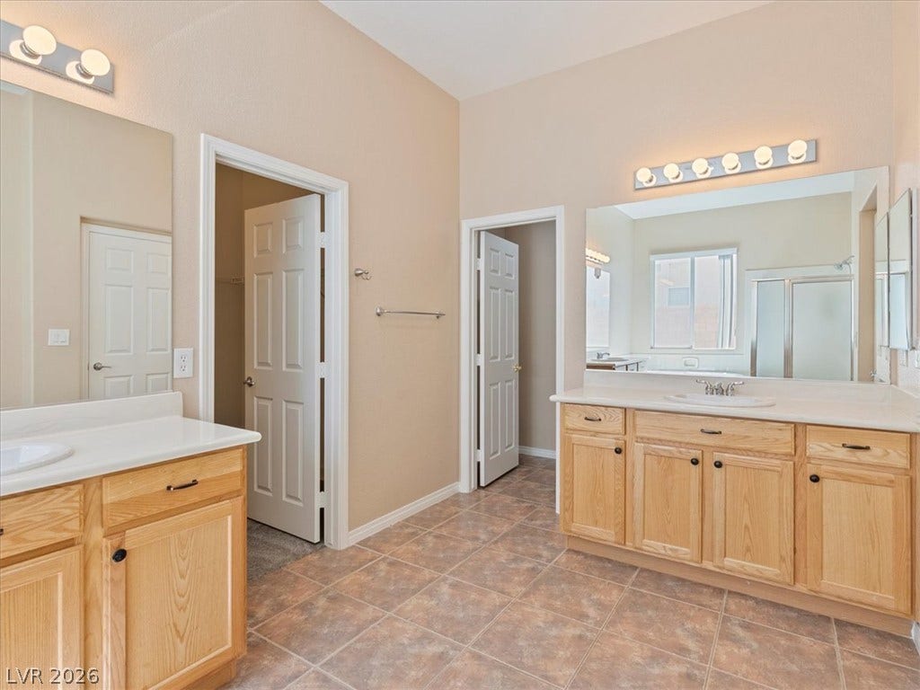 1217 Silent Sunset Avenue, North Las Vegas, NV 89084 A spacious layout showcases dual wood vanities and neutral tile flooring in this primary bathroom.