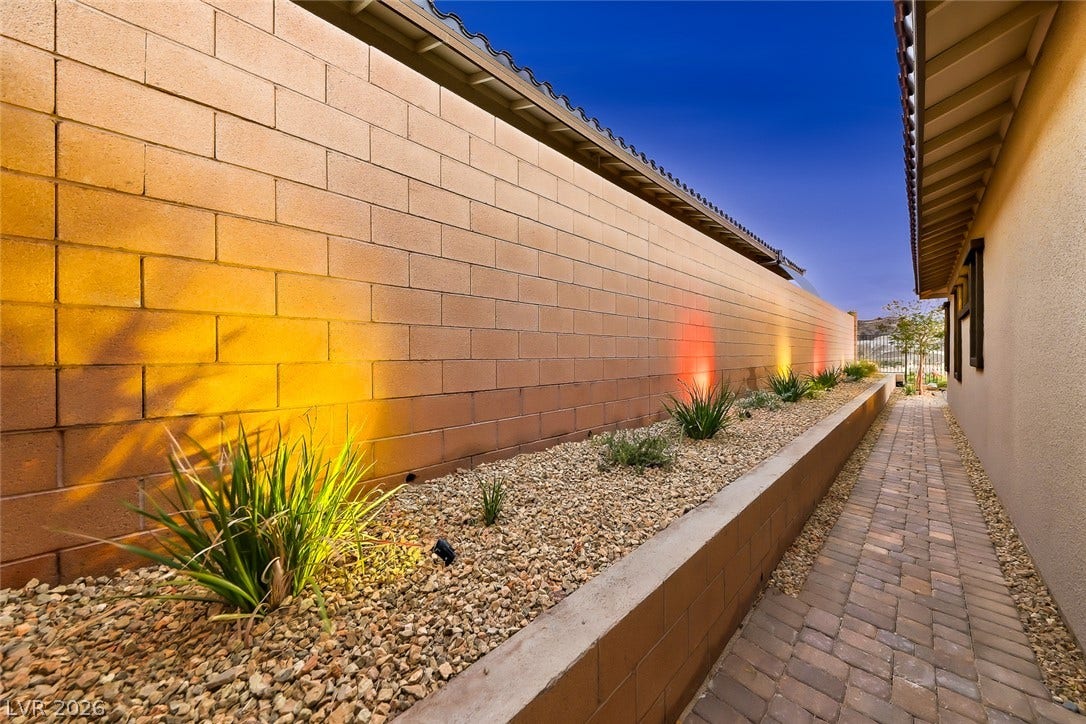 A paved side walkway runs alongside a raised stone garden bed with exterior uplighting.