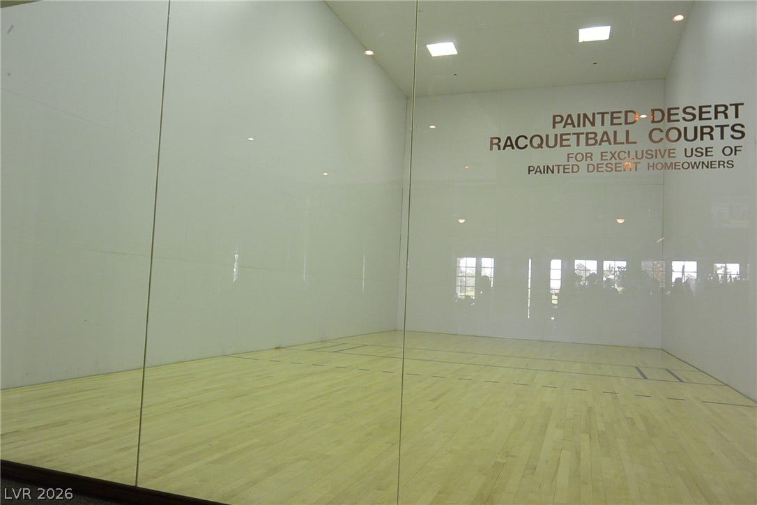 A glass-walled racquetball court features classic wood flooring and bright overhead lighting.