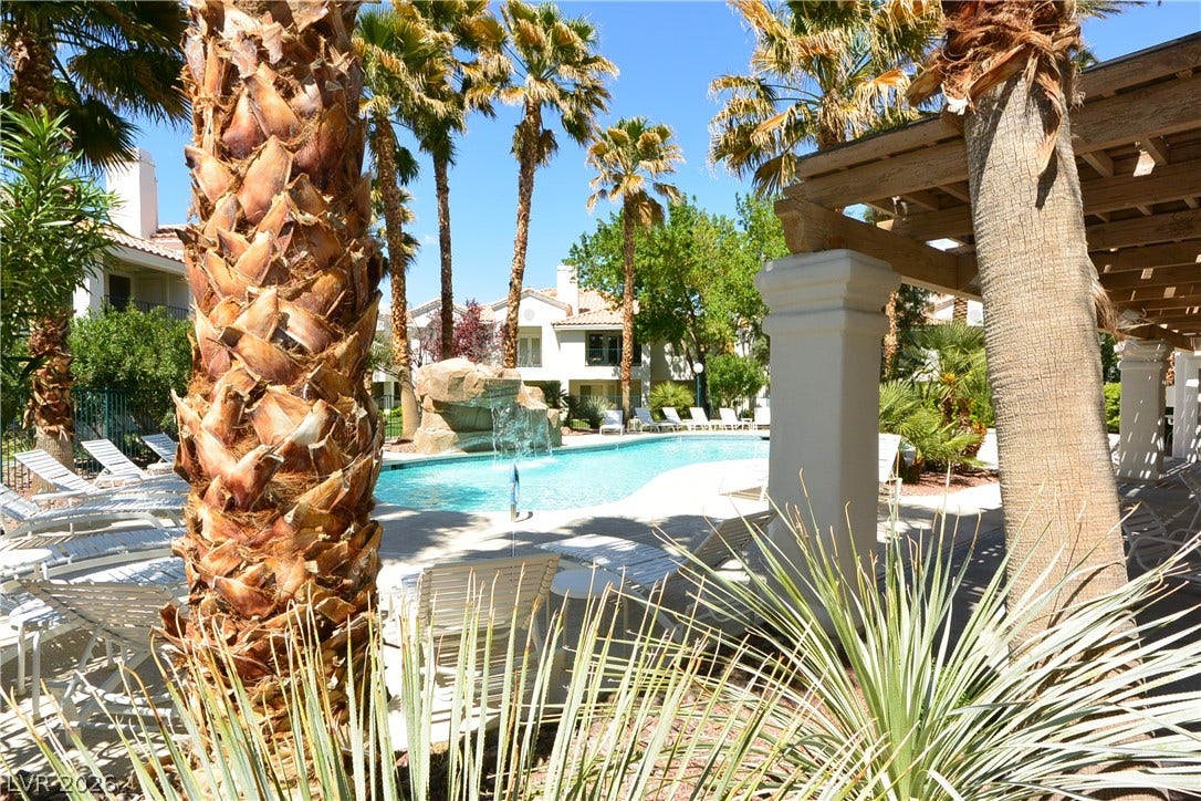 Towering palm trees and a shaded pergola frame a sparkling swimming pool with a rock waterfall.