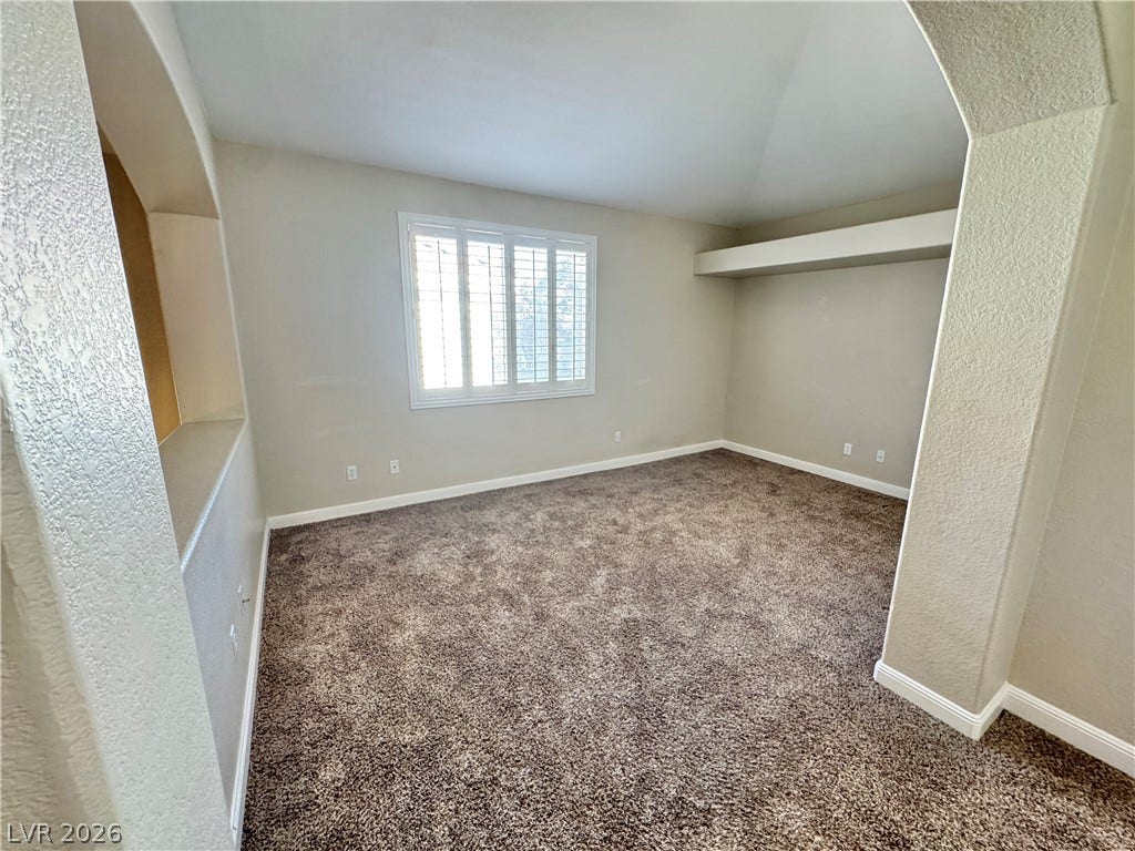 A spacious den/office features plush carpeting, unique alcoves, and a window with plantation shutters.
