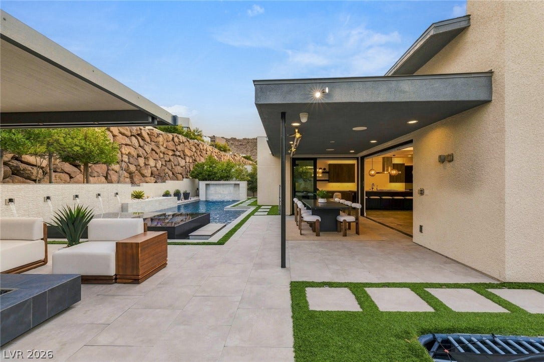 A spacious stone-paved patio adjoins a sleek swimming pool below a dramatic rock wall backdrop.