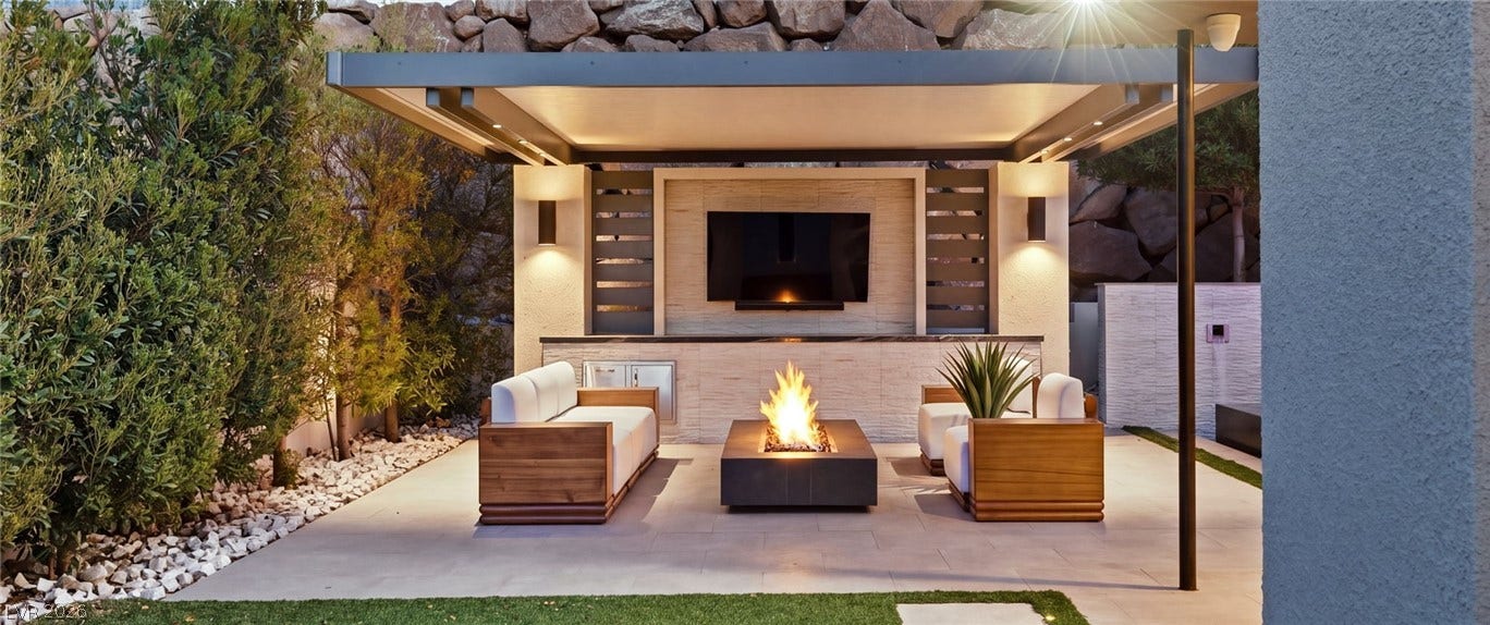 A stone patio features a modern fire pit sheltered beneath a contemporary covered structure.