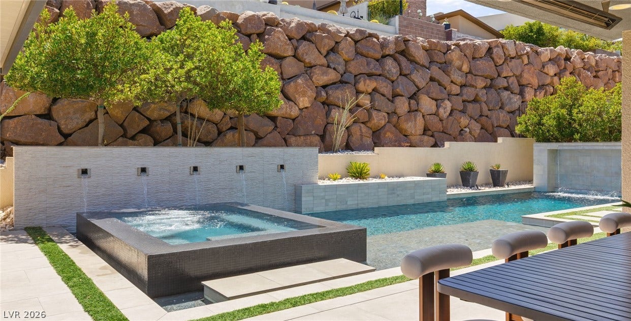 An engineered stone retaining wall towers behind a geometric pool, custom spa, and stone pavers.