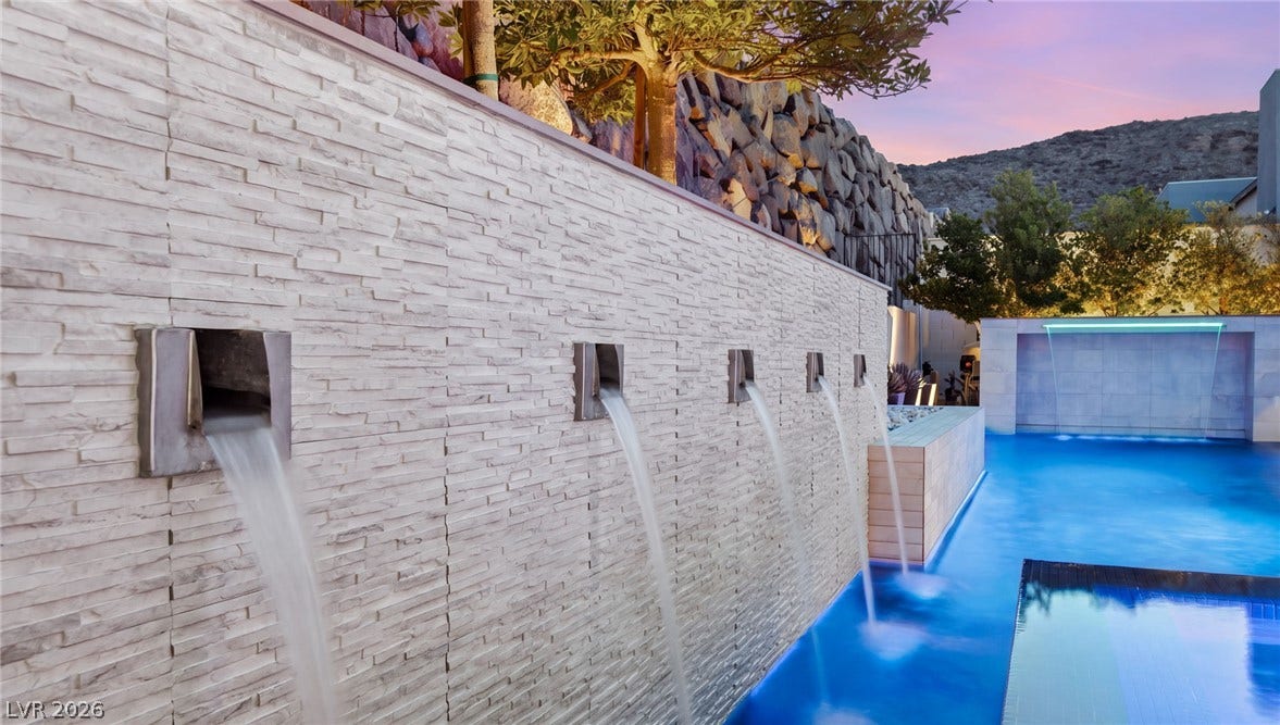 Custom stone water features pour directly into a shimmering backyard pool under twilight skies.