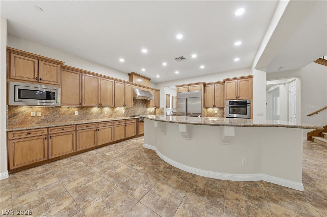 1357 Enchanted River Drive, Henderson, NV 89012 Ample wooden cabinetry and stone countertops frame a spacious tiled kitchen with a curved breakfast bar.